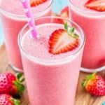 three strawberry smoothies in clear glasses with pink heart straws and strawberry garnish on a wooden board